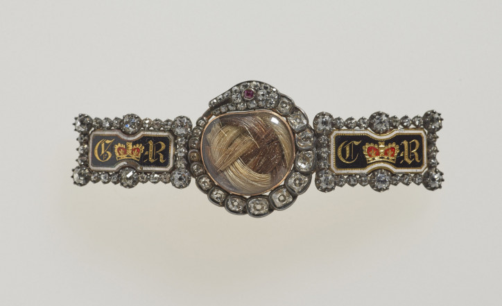 Zapięcie zawierające włosy króla Jerzego III i królowej Charlotty z XIX w., ozdobione diamentami i czarną emalią, z centralnym medalionem kryjącym skręcone kosmyki brązowych włosów monarchy oraz siwych jego małżonki, na którym widnieje połykający swój ogon diamentowy wąż z rubinowymi oczami. Po obu stronach emaliowane na czarno prostokątne panele ze złotymi monogramami GR i CR; zdjęcie: Royal Collection Trust/© Her Majesty Queen Elizabeth II 2021